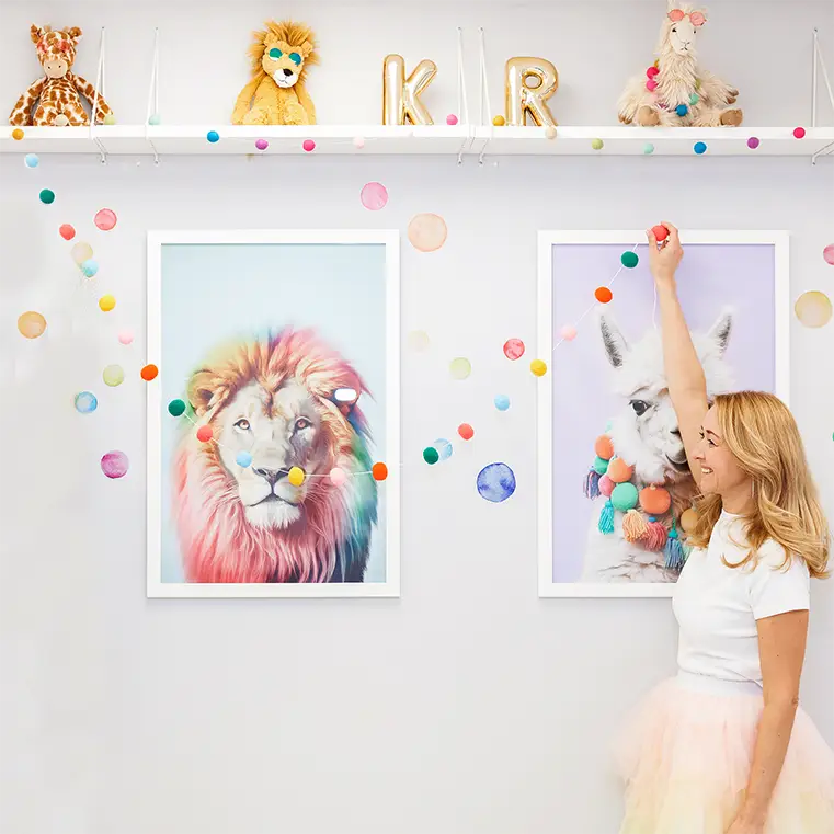 A woman holding up a pompom garland in front of a picture of a lion with a rainbow-coloured mane and a llama wearing a pompom necklace. On the shelf above them there is a toy giraffe, a toy lion, and a toy llama, all wearing toy sunglasses. In between the toys there are two gold ceramic letters spelling "KR".