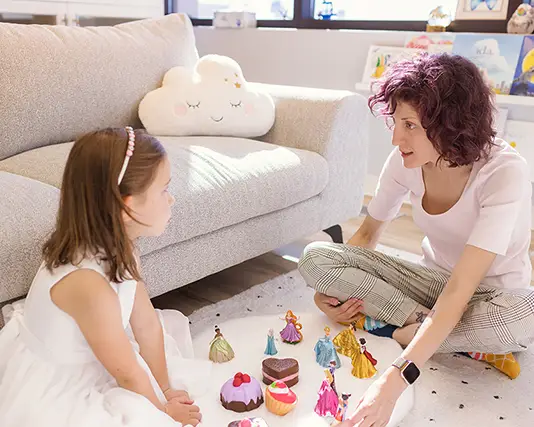 A girl and a therapist doing play therapy in a specialized child counselling session. They are sitting on a white carpet, playing with princess toys.