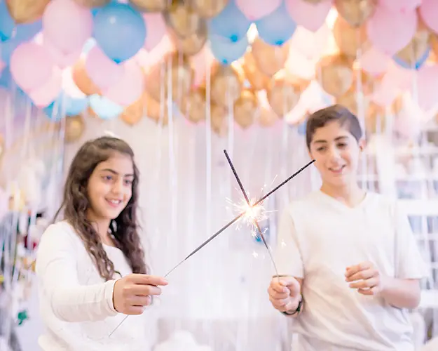 A girl and boy with sparklers. Dozens of blue, gold, and pink helium balloons float overhead in a specialized child counselling group session.