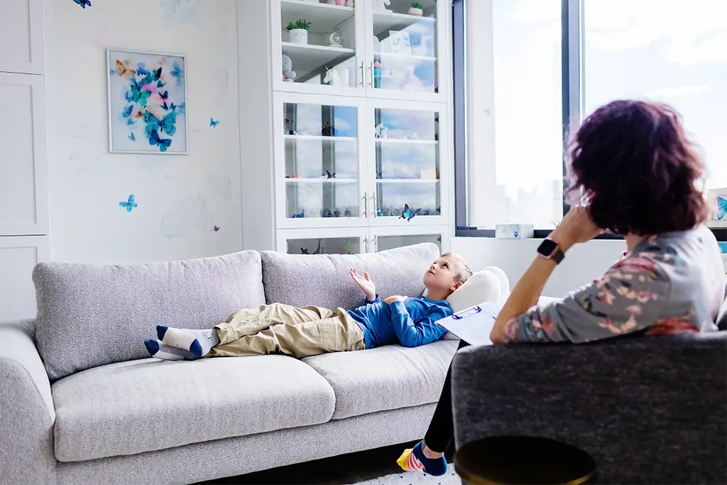 Child and counsellor in a playroom with a blue butterfly. The boy is lying on a sofa and holding out his hand as the butterfly flies above his head. The child counsellor is in the foreground.