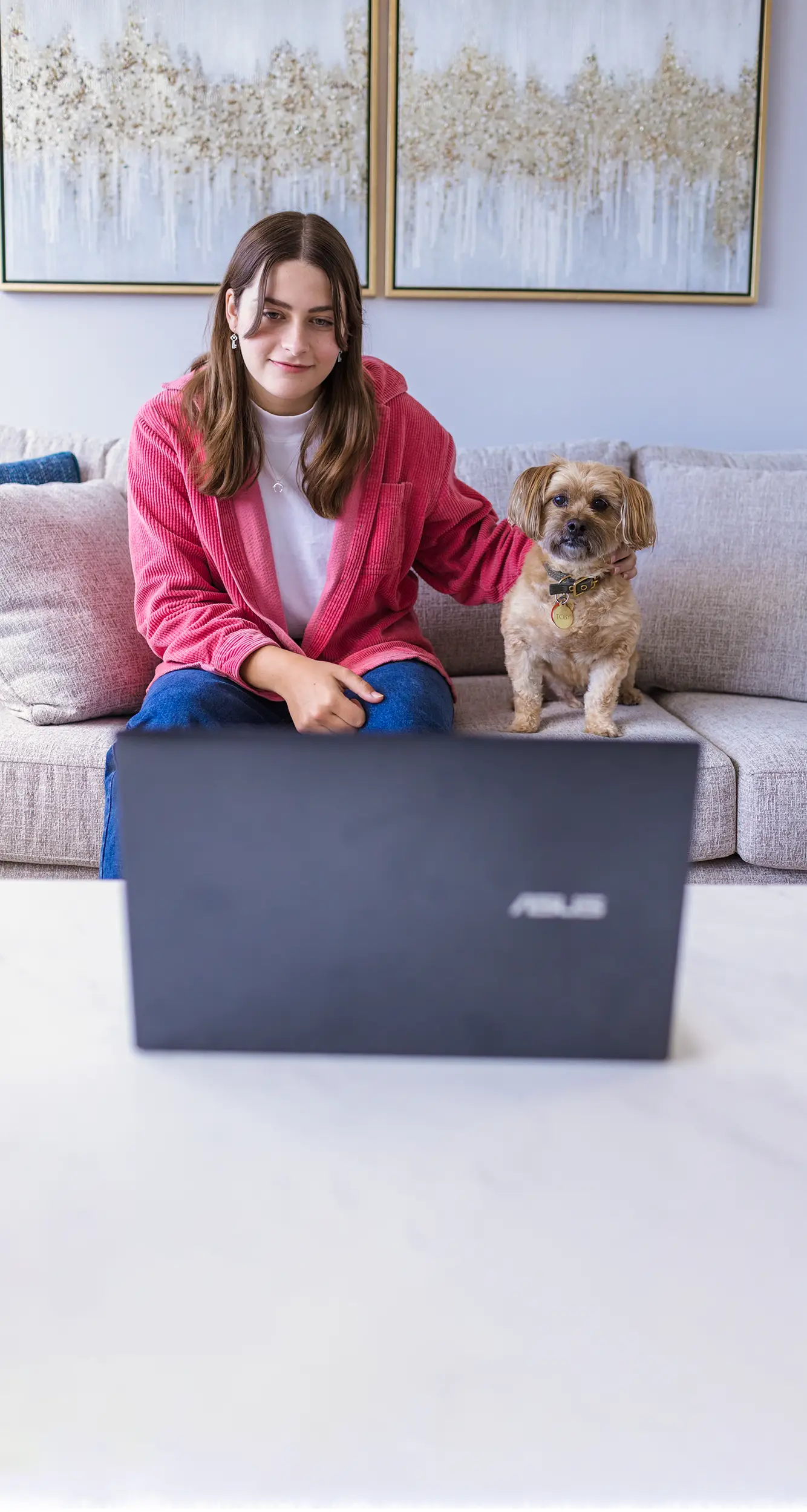 An adolescent girl sitting on a couch, with her small dog next to her, with a laptop in front of them. They are in a telepsychology (online therapy) session.