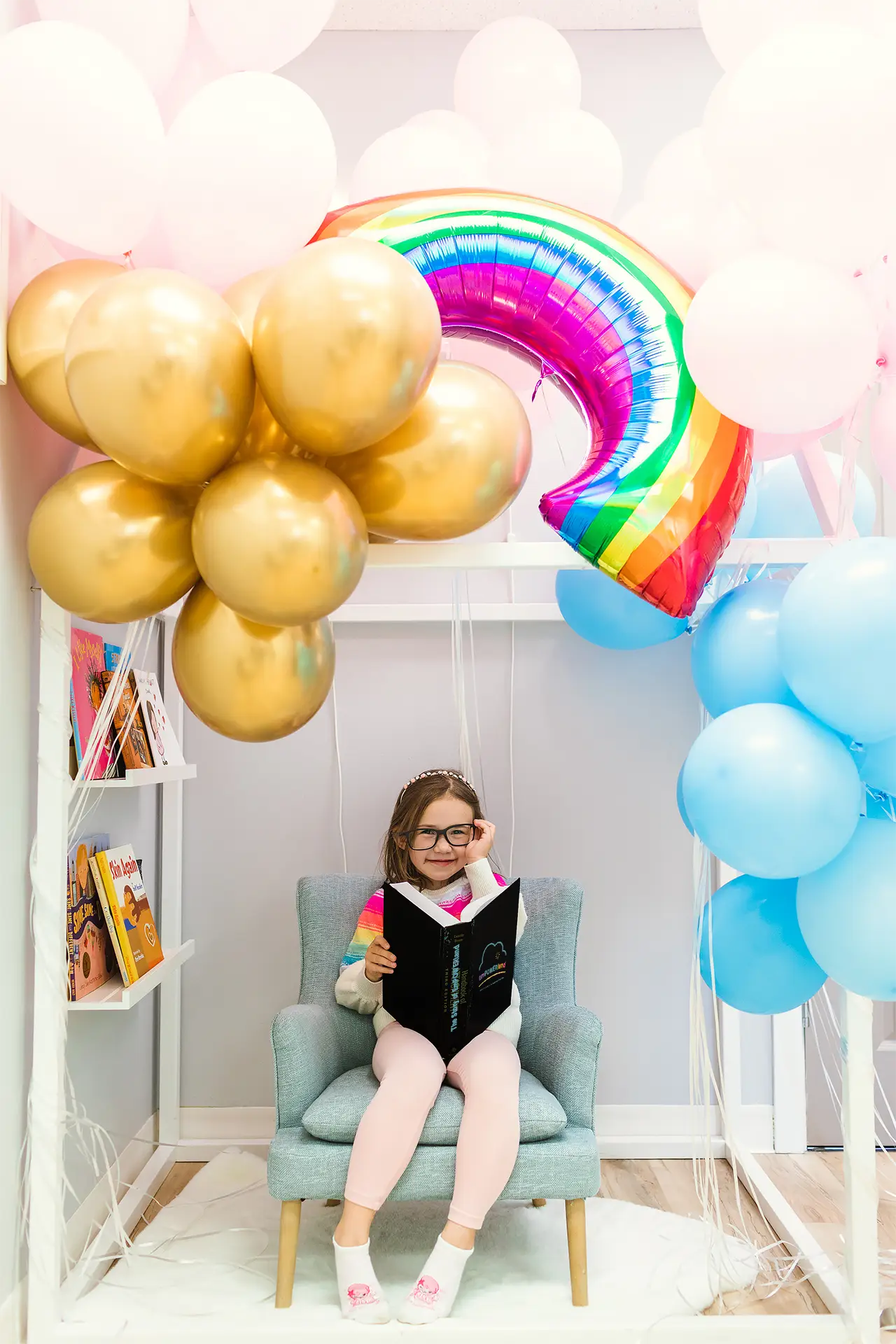 A child smiles as she reads The Story of EmPOWERland. She is wearing toy glasses. Gold, pink, blue, and rainbow balloons shine overhead.