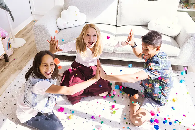 A girl and a boy sitting cross-legged on a rug in a play therapy session with a play therapist give each other high-fives. They are surrounded by multicoloured pompoms.