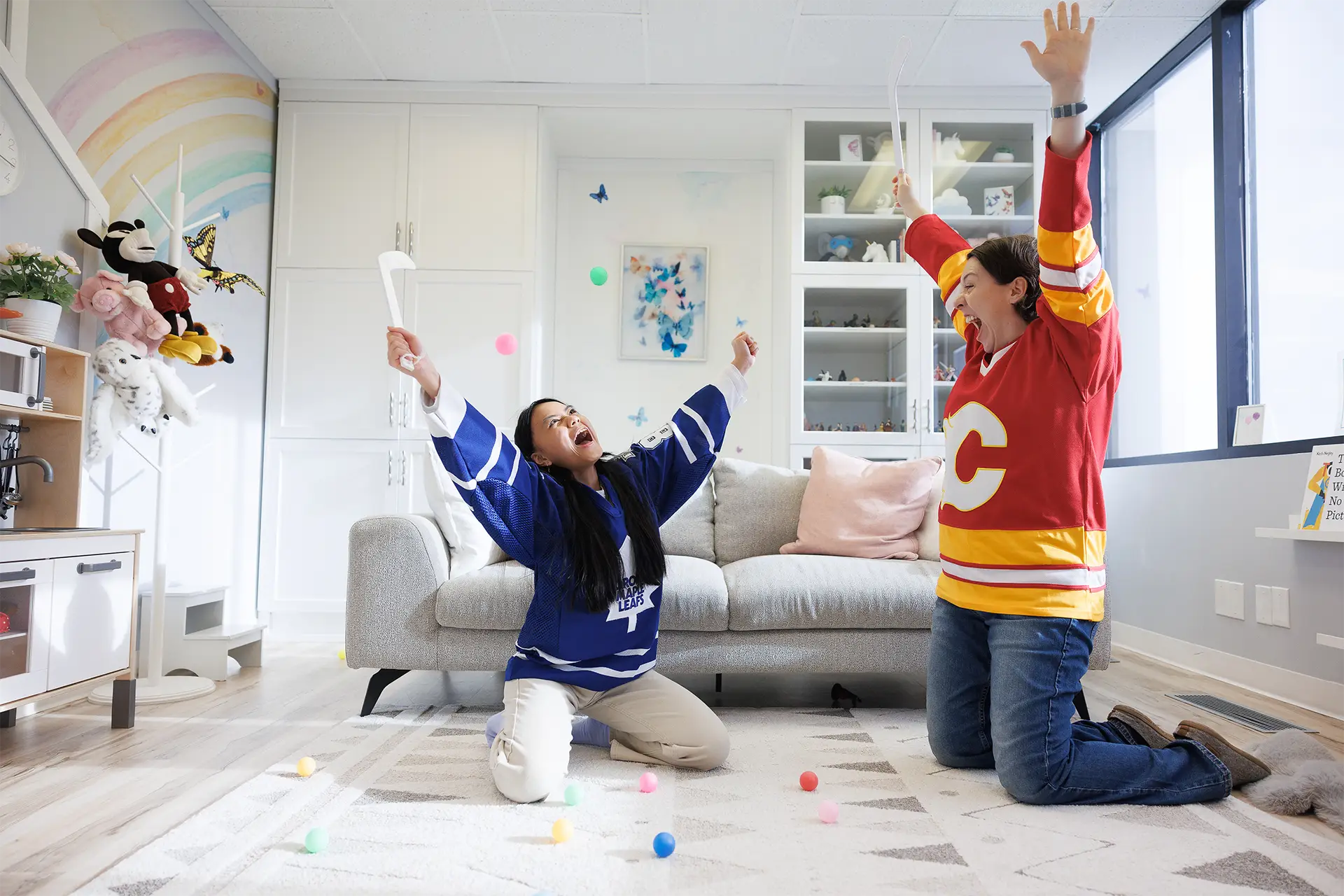 Calgary child therapist Chelsea Houston with a teen in a play therapy session at Kids Reconnect. Both the child and the teen are smiling and cheering as they hold their hands up in the air in celebration, holding a small toy hockey stick. Chelsea is wearing a red Calgary Flames hockey jersey, and the girl is wearing a blue Maple Leafs hockey jersey. There are coloured balls around them and flying in the air.