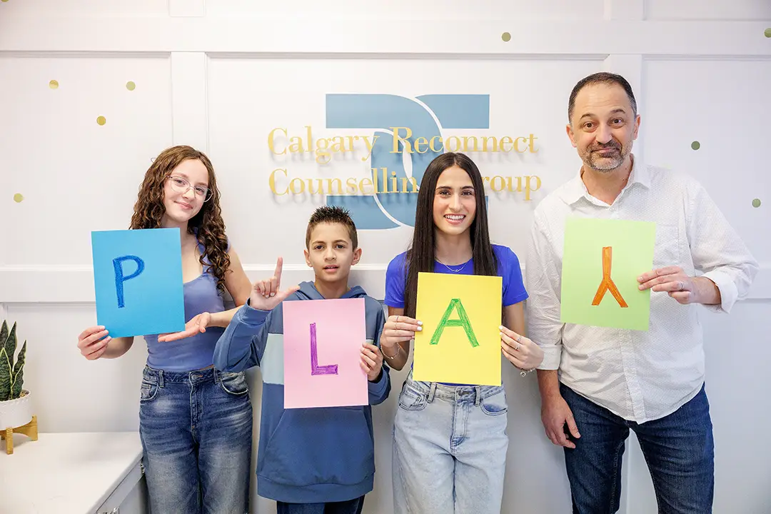 Two girls, a boy, and a Dad standing in front of the Calgary Reconnect Counselling Group sign. Each is holding a sign with a letter to spell PLAY. The Dad is playfully holding the "Y" upside down and making a funny face. Everyone shows a different PLAY style!