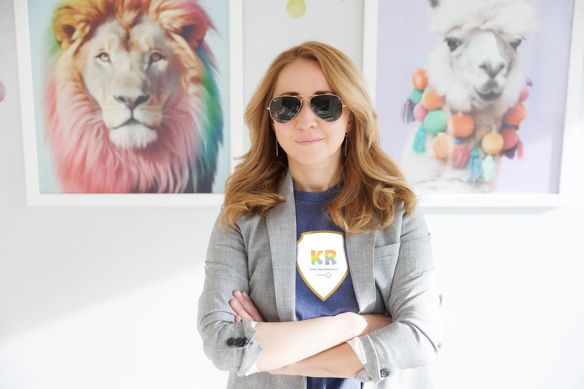 Natalie Bergman wearing a Kids Reconnect t-shirt Child therapist Natalie Bergman wearing sunglasses and a Kids Reconnect under a suit jacket with rolled-up sleeves. Behind her are two pictures: on the left, a lion with a rainbow-coloured mane, and on the right, a llama wearing a rainbow-coloured pompom necklace