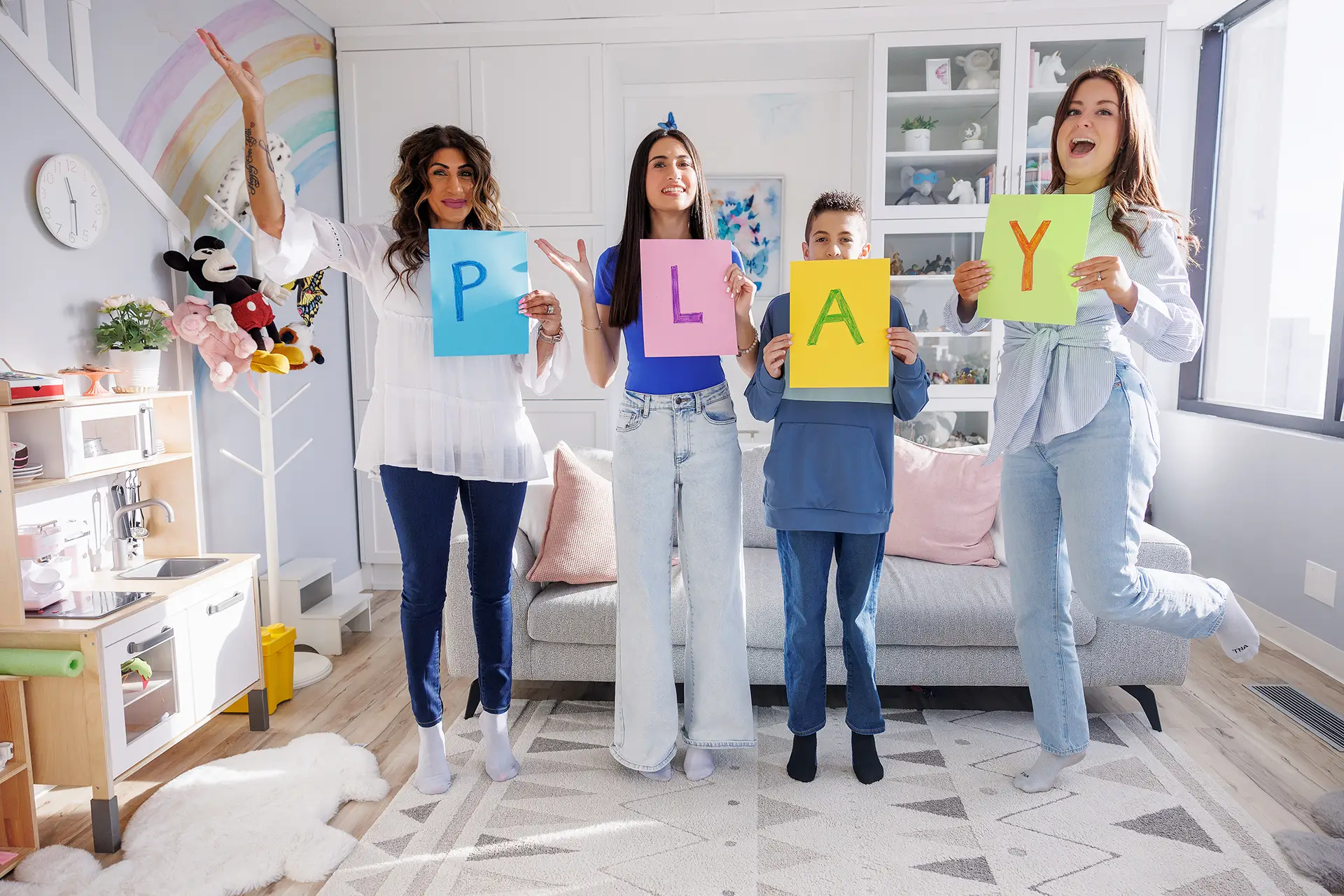 A mom, am adolescent gir, a boy, and a child psychologist stand in a playroom as they each hold a coloured sign with one letter. Together, they spell the word P-L-A-Y!