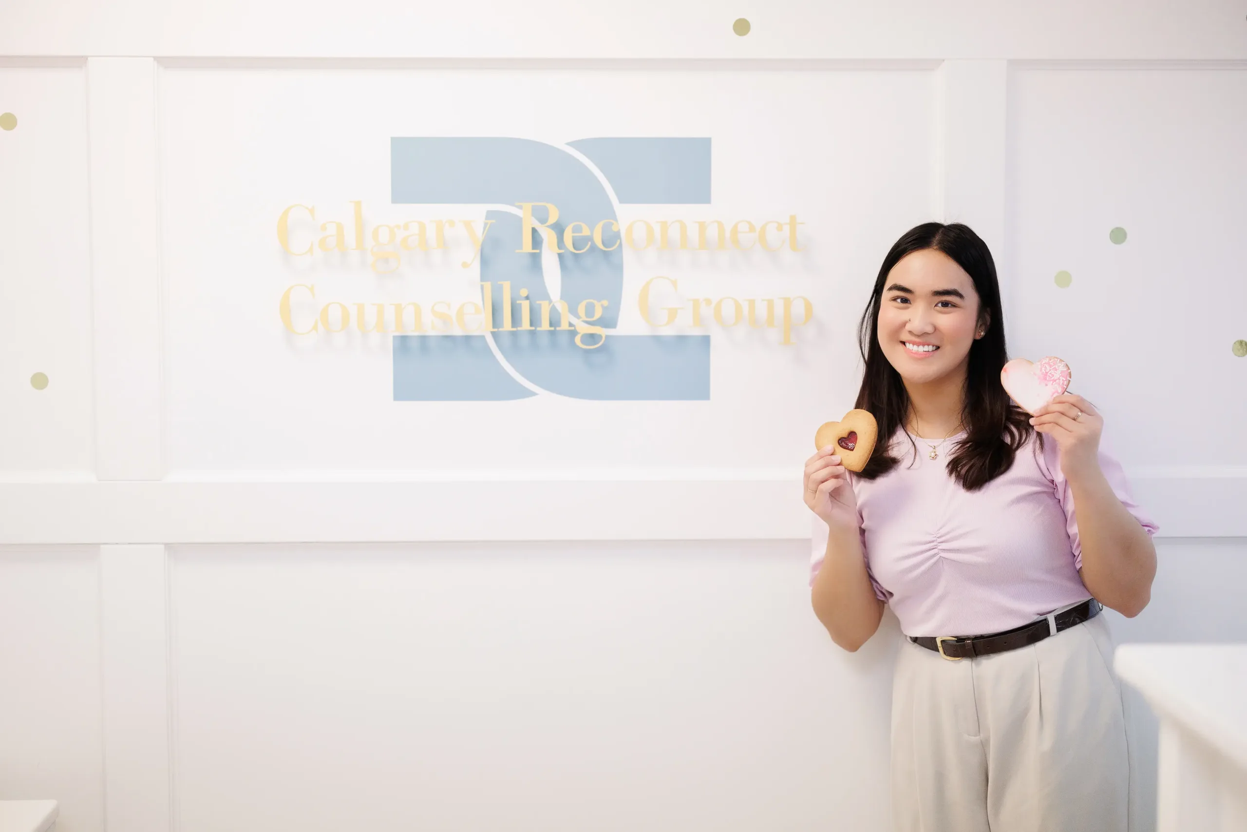 Our friendly Intake Team member Mary Molina holds two heart-shaped cookies as she stands in front of the Calgary Reconnect Counsellig Group sign at the reception area.