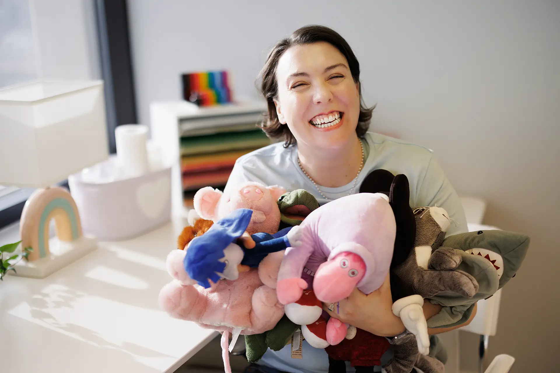 A child therapists smiles widely as she holds a dozen stuffies in her arms in a playroom at Kids Reconnect. It's Chelsea Houston, Superhero child therapist!