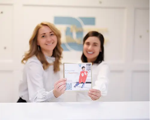 Calgary child psychologists Natalie Bergman and Marcella Galizia holding a gold framed card of a boy wearing a red Superhero cape. The text in the card reads "Every child is a Superhero".