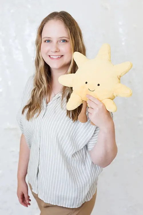 A woman holding a smiling sun stuffy toy. Headshot of Kate Holmberg, Child Therspist at Kids Reconnect.