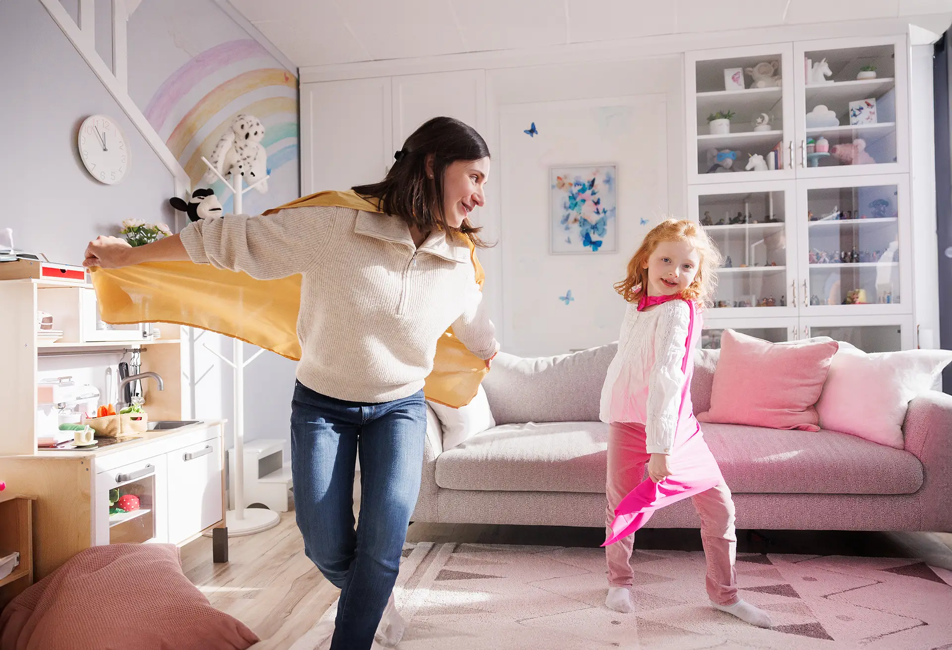 Play therapy for Superhero kids at Kids Reconnect in Calgary A woman and a girl swirling their Superhero capes in a playroom with a rainbow and butterfly murals, a toy kitchen, and a sofa in the background.