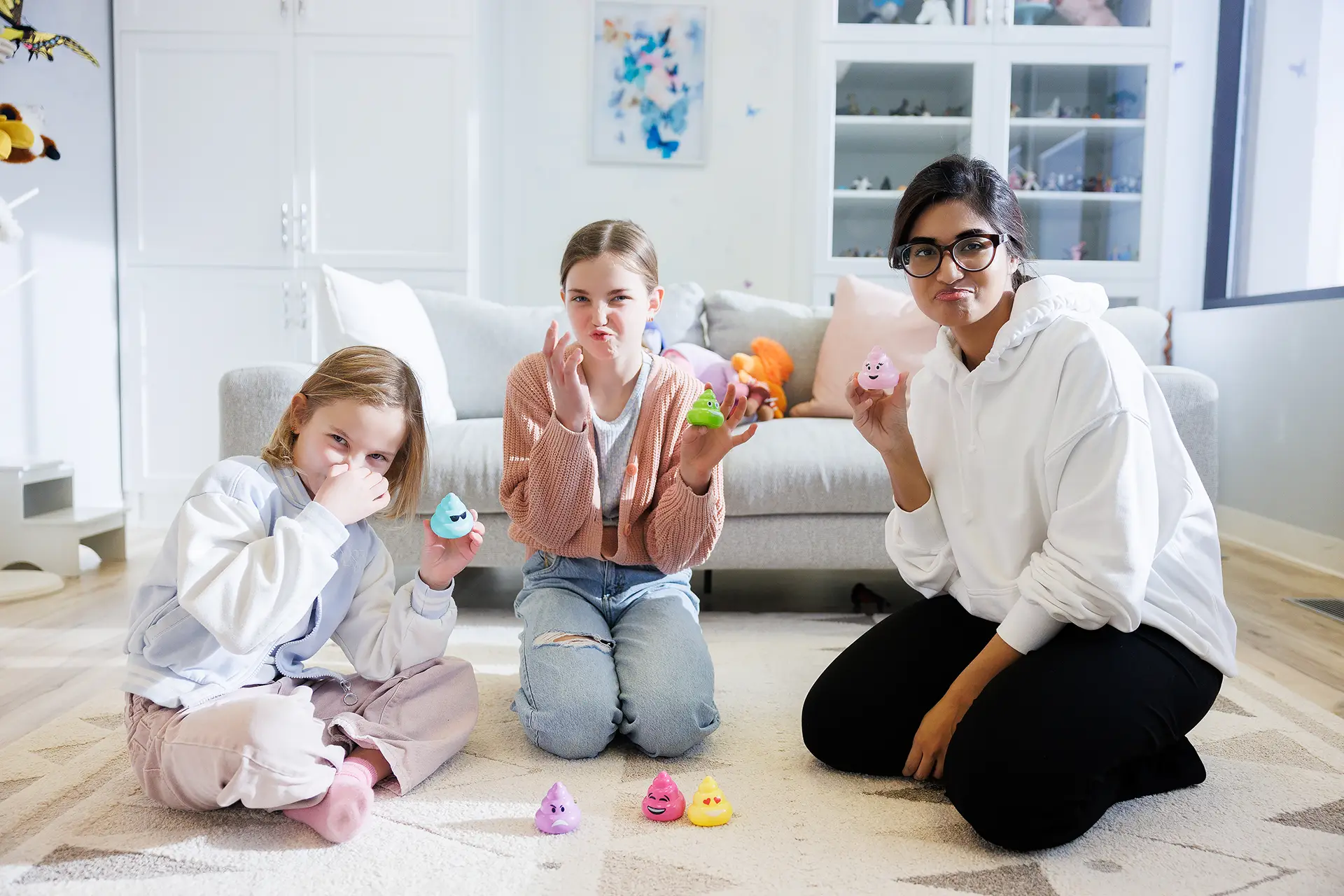 A woman and two girls sitting on the rug in a playroom, wrinkling and pinching their nose playfully as they hod up "poop" emoji toys in rainbow colours.